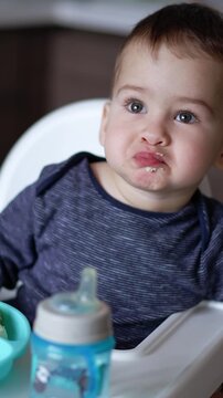Cute toddler boy sits in high chair leaned to the side. Baby is focused on something while mother feeds him. Blurred backdrop. Vertical video