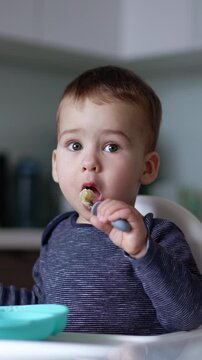 Caucasian toddler learning to use a form. Beautiful baby boy eating himself sitting at high chair. Blurred backdrop. Vertical video