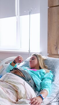 Pregnant blonde Caucasian woman lies in the hospital bed connected to equipment. Cardiotocography belt is on the belly of patient. Vertical video
