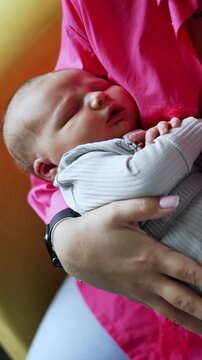 Unrecognized woman in pink shirt holding her baby. Mother sits in an armchair rocking her newborn child in blue romper. Vertical video