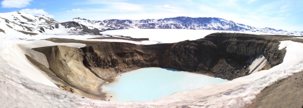 Panoramic view of a volcanic crater lake surrounded by rugged cliffs and snow-covered mountains,