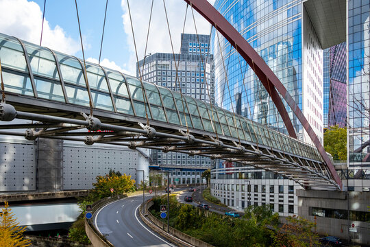 Highway traffic passes under a modern bridge with architecture framing a corporate business finance skyline of glass towers at La Defense Paris urban connection