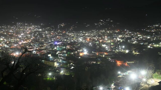 Wide panoramic view of a city illuminated at night with glowing lights across buildings and streets. Captured from an elevated viewpoint, ideal for timelapse, urban life, nightlife, and cityscape conc