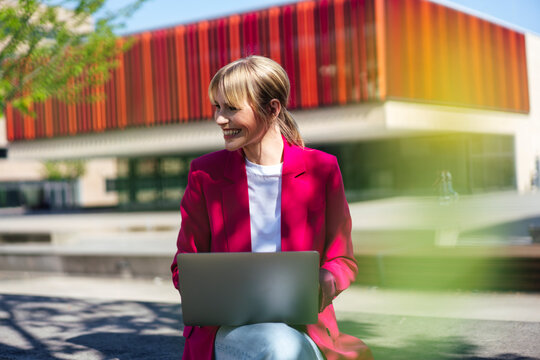 Happy blond businesswoman with laptop sitting at office park under sunlight