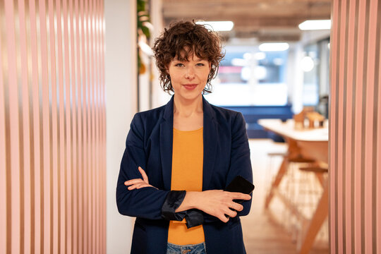 Young businesswoman holding smart phone and standing with arms crossed in office