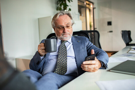 Senior businessman checking smartphone while drinking coffee in office
