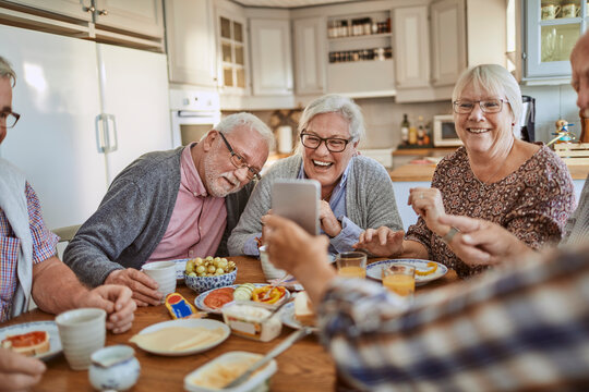 Senior friends laughing at smartphone during breakfast in home kitchen