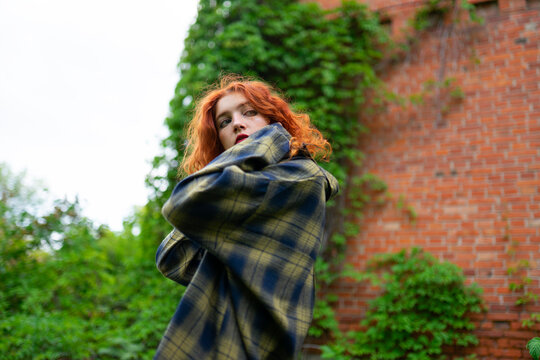 Woman with red curly hair in casual jacket outdoors by brick wall