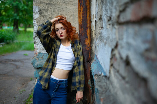 Red haired woman in casual jeans leaning by old urban wall outdoors