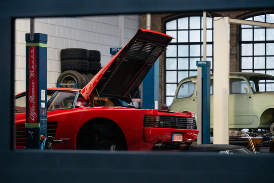 red Ferrari Testarossa with opened hood in auto workshop. Rear end of 1980s sportscar on hydraulic lift inside auto repair shop with other old cars in the background. Berlin, Germany - January 14 2026