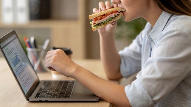 Multitasking woman in shirt pointing at laptop chart biting sandwich typing at desk, copy space