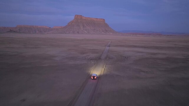 Aerial view of a car with headlights on driving along a dirt road towards Factory Butte in the desert at twilight Wayne County, Utah, United States.