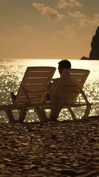 Man beach sunset, relaxing on a chair by the ocean during golden hour, tranquil vacation, vertical video