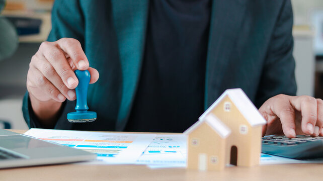 Close-up of business person stamping approval on real estate documents with house model, calculator, and laptop. Concept of mortgage loan, property investment, and financial decision.