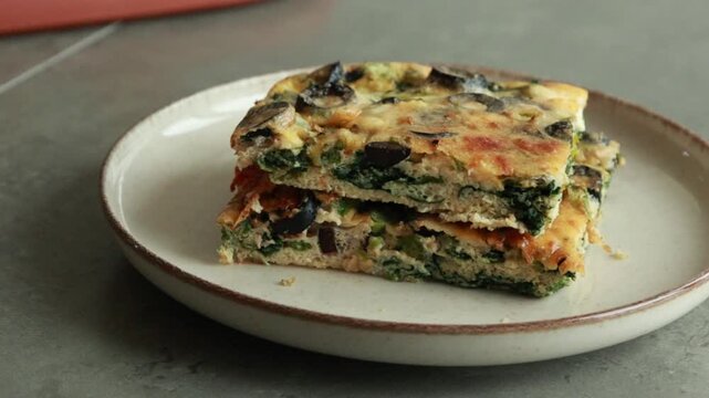 Close-up of a slice of baked egg frittata with olives and spinach on a rustic plate