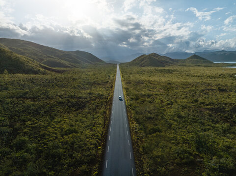 Aerial view of a solitary car slicing through a verdant landscape, the road a dark ribbon bisecting the bright green expanse under a sky of dramatic clouds, Goro, South Province, New Caledonia.