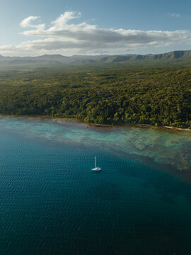Aerial view of a lone sailboat gliding on the turquoise waters where the deep blue sea meets the lush green forest, Goro, South Province, New Caledonia.