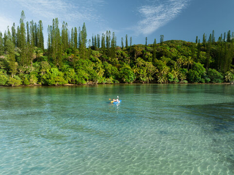 Aerial view of a two kayakers drifting on crystal-clear waters, embraced by the verdant coastline and towering pines, Goro, South Province, New Caledonia.
