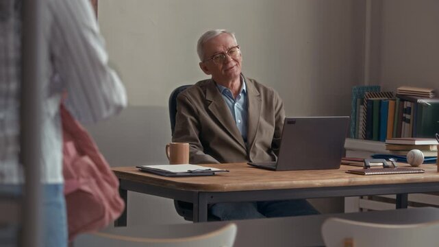 Stab medium eye level shot of senior Caucasian male teacher sitting at desk greeting students arriving in classroom and taking books out of backpacks on desks, beginning lesson