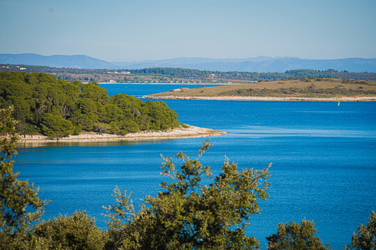 View of the tranquil turquoise sea meeting the rugged coastline, verdant trees, and distant mountains under a clear sky, Pula, Istria County, Croatia.