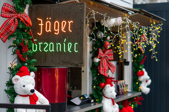 Mulled wine stall at a Christmas market with Jager Grzaniec neon sign, festive decorations featuring white teddy bears, garlands and LED lights in Poznan, winter holiday atmosphere.