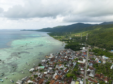 Aerial view of the coastal village nestled between the turquoise sea and verdant hills under a cloudy sky, Karimunjawa, Central Java, Indonesia.