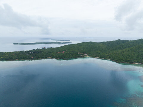 Aerial view of the island's lush green canopy meeting the turquoise sea, a tropical haven under a soft, cloudy sky, Karimunjawa, Central Java, Indonesia.