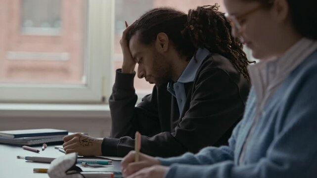 Side view waist up of Biracial male college student with dreadlocks concentrating on notes while sitting at desk during lesson with classmate writing nearby