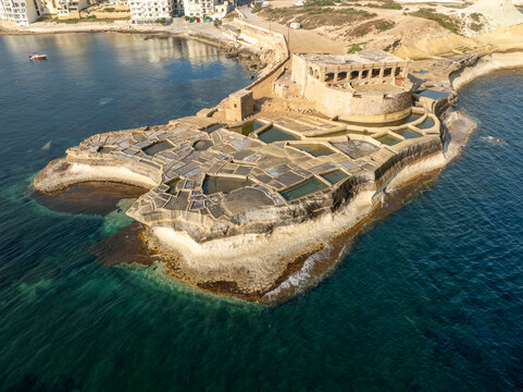 Aerial view of the geometric salt pans creating a textured mosaic against the turquoise sea near the historic Qolla l-Bajda Battery, Iz-Zebbug, Gozo, Malta.