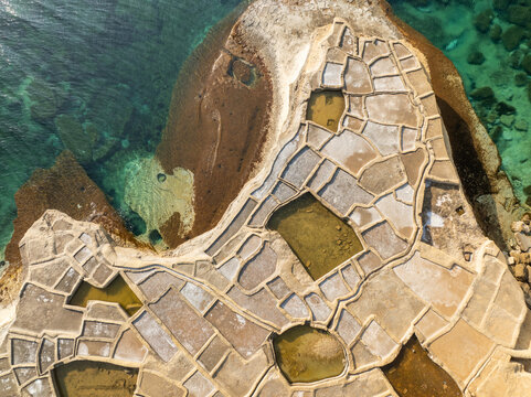 Aerial view of the Qolla I-Bajda Salt Pans carved into the rocky coastline creating a geometric pattern against the turquoise sea, Iz-Zebbug, Malta.