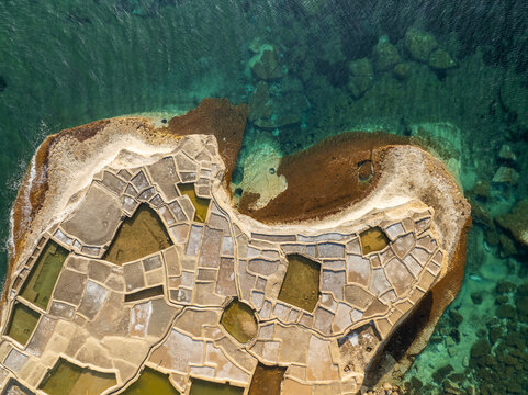 Aerial view of the ancient Qolla I-Bajda Salt Pans shimmer under the summer sun, with turquoise waters contrasting against the geometric patterns, Iz-Zebbug, Malta.
