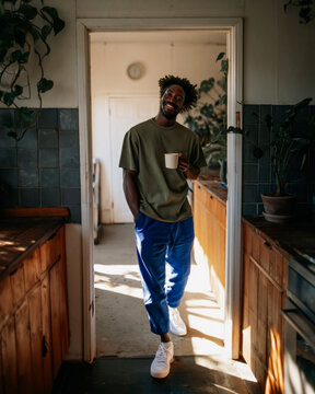 Smiling Young Man Leaning Against Wooden Cabinet in Sunlit Modern Apartment Wearing Casual Green T-Shirt and Navy Trousers
