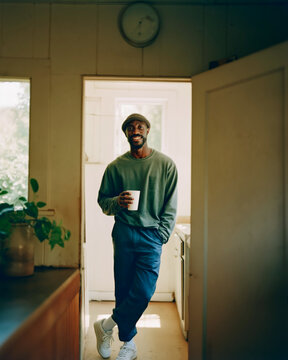 Smiling Young Man Leaning Against Wooden Cabinet in Sunlit Modern Apartment Wearing Casual Green T-Shirt and Navy Trousers