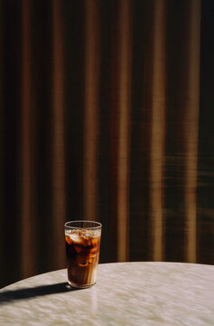 Glass of Cola With Ice Cubes on Wooden Table in Warm Sunlight With Dramatic Shadows and Soft Curtain Background
