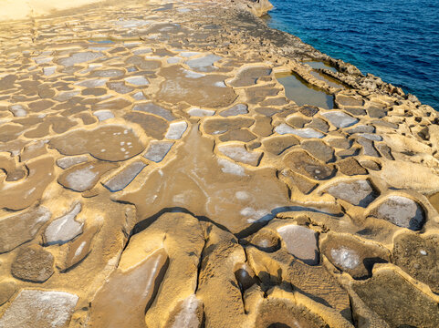Aerial view of rugged, sun-baked salt pans shimmer with residual water, contrasting sharply with the deep blue sea, Iz-Zebbug, Gozo, Malta.