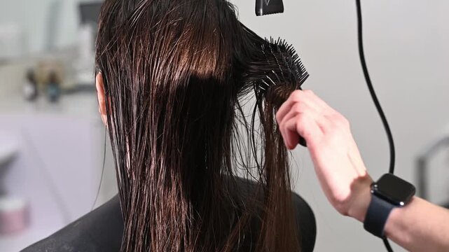 A hairdresser blow-dries a client's long, natural, dark hair with a round brush and a styler at a salon. Professional hair styling and care at the hair salon.