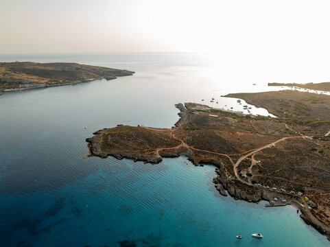 Aerial view of turquoise waters meeting rugged coastline, boats dotting the horizon under a hazy sky, Blue lagoon, Comino, Malta.