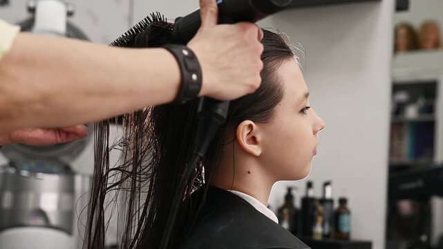 A hairdresser blow-dries a client's long, natural, dark hair with a round brush and a styler at a salon. Professional hair styling and care at the hair salon.