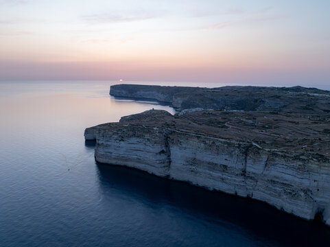 Aerial view of rugged cliffs meeting the tranquil sea under a soft, pastel sky at Sanap Cliffs, Munxar, Gozo, Malta.