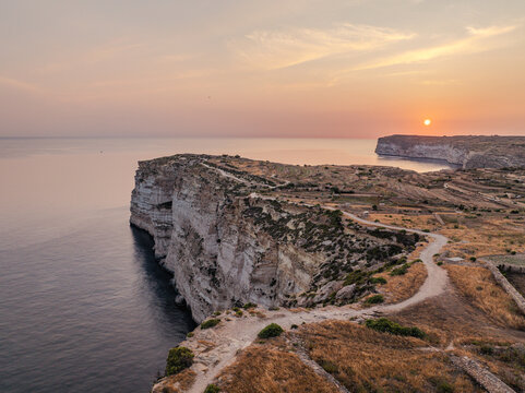 Aerial view of rugged, sun-kissed cliffs meeting the tranquil, dark blue sea under a pastel sky at Sanap Cliffs, Munxar, Gozo, Malta.