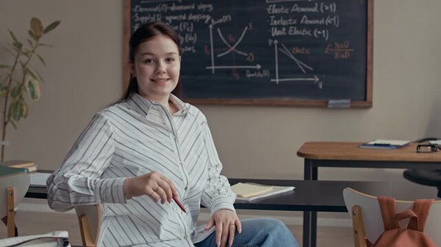 Medium portrait of Caucasian female student smiling at camera while sitting at classroom desk against blackboard with math theory