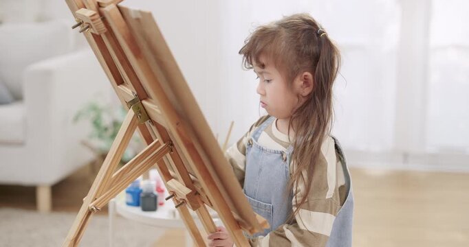 Asian toddler daughter stands beside wooden easel, paints canvas with small brush, keeps serious face, repeats stroke practice, builds discipline, perseverance, creative learning near bright window.