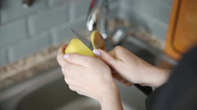Close up hands peeling fresh yellow potato over kitchen sink with grey tiled background