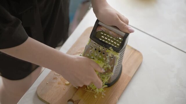 Hands grating fresh zucchini over wooden cutting board in bright kitchen environment