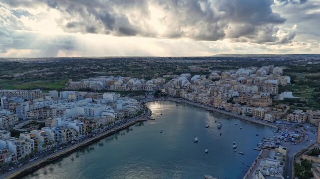 Marsaskala, Malta, a coastal town with docked boats along the bay, Mediterranean residential architecture, streets, and waterfront under a dramatic, cloudy sky in an aerial hyperlapse view.