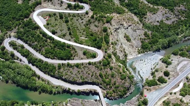 Cinematic drone shot of a scenic winding road with sharp hairpin turns. High-angle view showing the zig-zag route over the Venetikos river and lush green cliffs in Grevena. Ideal for travel films.