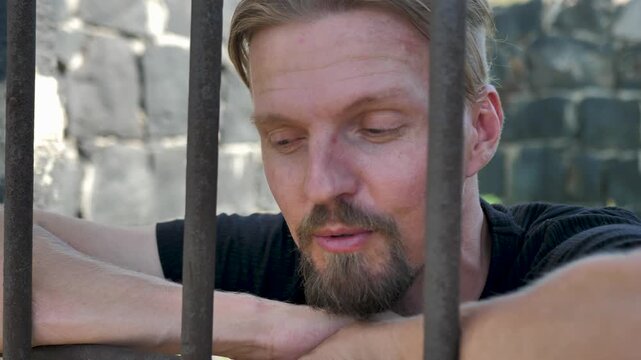 Young caucasian prisoner behind iron bars in good mood smiling and smirking without remorse for committed crime. Bearded criminal in prison. Recidivist and illegal migrant