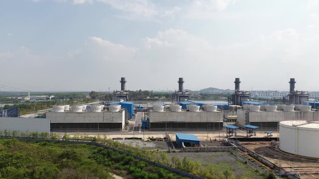 Ascending aerial view of a large combined cycle gas turbine power station, showcasing its cooling towers, smokestacks, and surrounding industrial infrastructure under a clear sky