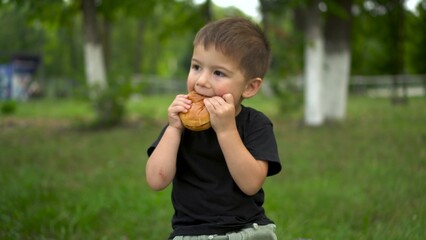 Fototapeta premium Three year old boy eats cheeseburger outdoors and looks at camera portrait 4k