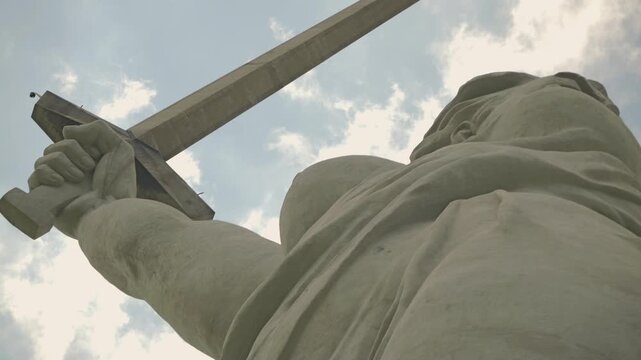 Imposing low angle shot of a massive concrete monument depicting a woman with a raised sword. The dramatic perspective emphasizes her power scale against a soft, cloudy sky. Motherland calls